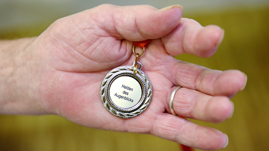 Eine Hand hält eine Medaille fest, auf der "Helden des Augenblicks" steht. 