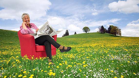 Eine Dame, die in einem roten Sessel auf einer Blumewiese sitzt und Zeitung liest