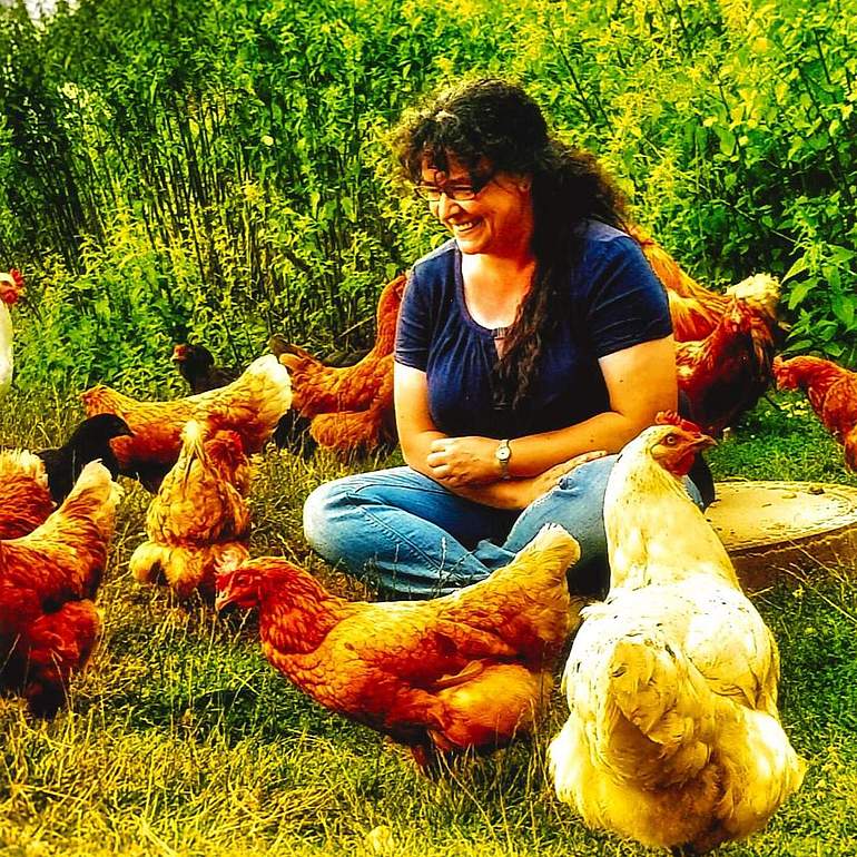 Angela Panico sitzt im Schneidersitzt auf einer Wiese und ist umgeben von Hühnern.