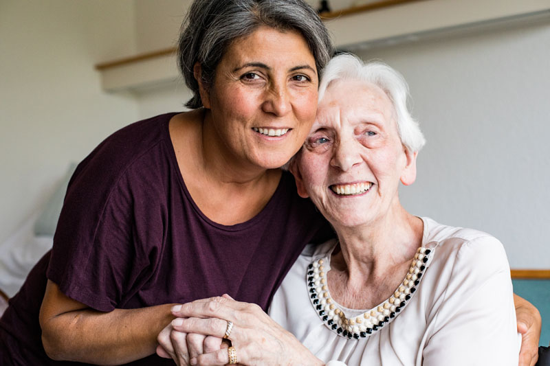 Zierliche Frau mit dunkelen kurzen Haaren umarmt alte, grauhaarige Frau. Beide lächeln.