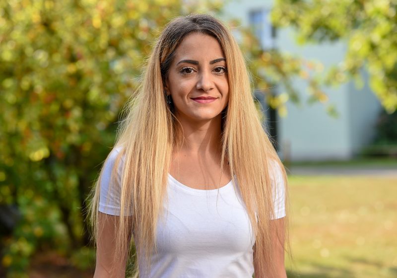 Frau steht draußen unter Herbstbäumen. Lange blonde Haare, weißes T-Shirt.