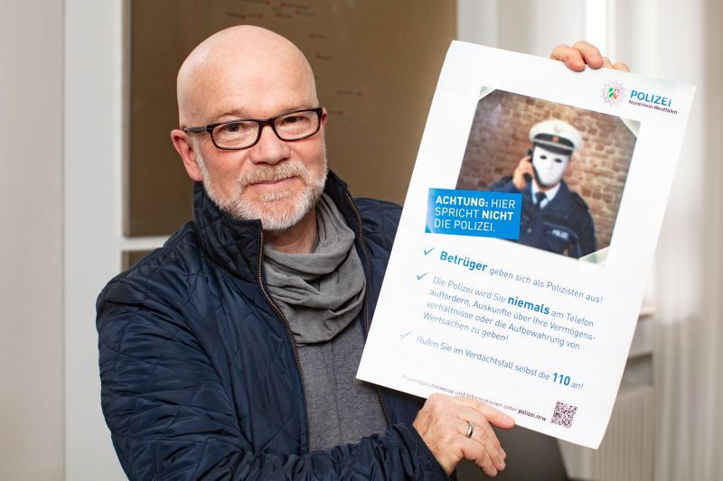 Porträt von Mann mit Brille, weißem Bart und Glatze, der ein Plakat mit Warnung vor Betrügern ins Bild hält