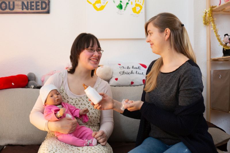 Frau hält einen Babysimulator auf dem Arm und unterhält sich mit einer anderen Frau.