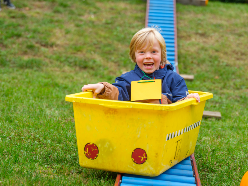 Ein Junge sitzt in einer gelben Kiste auf einer Rollenbahn.