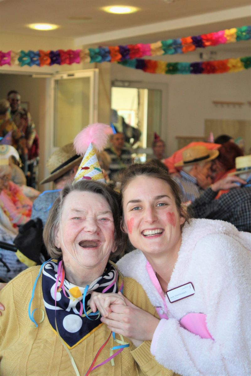 Betreuerin im Hasenkostüm umarmt Bewohnerin mit Partyhütchen, beide lachen in die Kamera