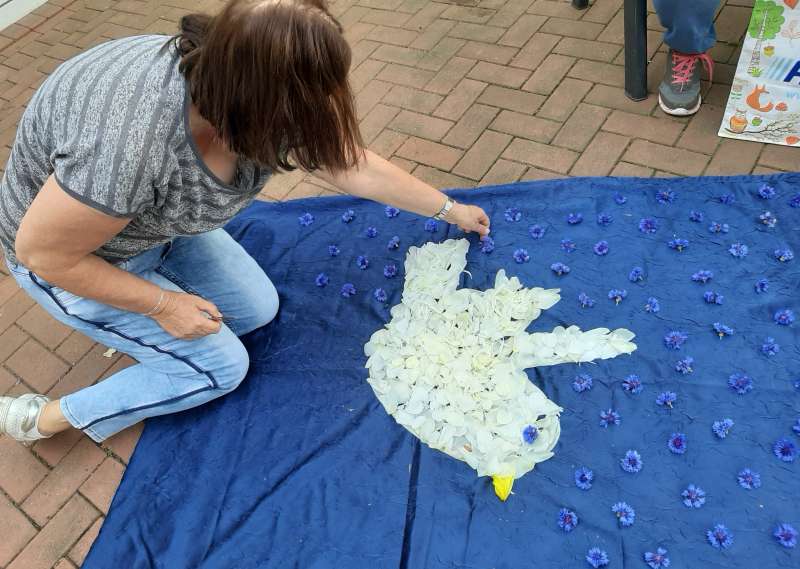 Gottesdienst der besonderen Art: eine Ehrenamtliche schmückt den Innenhof des Käthe-Kollwitz-Hauses mit einem Blumenteppich, der eine weiße Taube auf blauem Tuch zeigt.