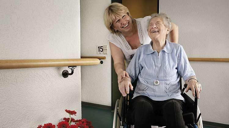 Gut betreut im Johanneswerk: Pflegekraft begleitet lachende ältere Dame im Rollstuhl