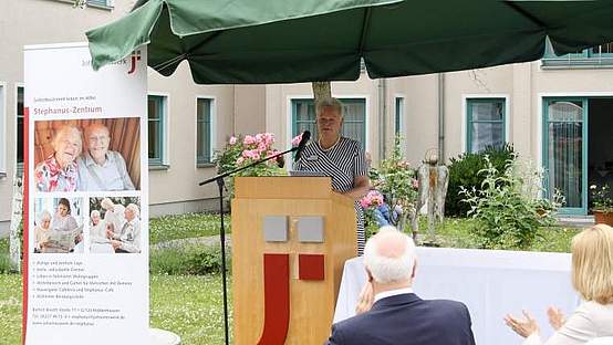 Hausleiterin Manuela Schock steht draußen an einem Rednerpult unter einem Sonnenschirm. Daneben steht ein Banner mit dem neuen Namen Stephanus-Zentrum.