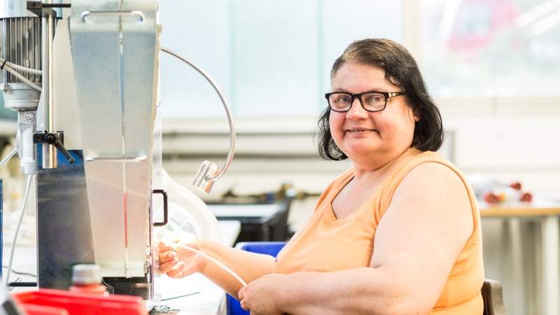 Frau mit braunen Haaren und orangem Top sitzt vor einer Maschine und hält ein kabel in den Händen