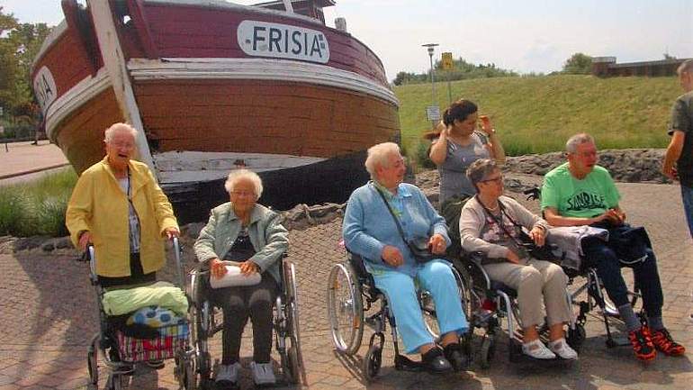 Sechs Menschen, davon vier im Rollstuhl und einer mit Rollator, vor einem alten Schiff, das an Land liegt