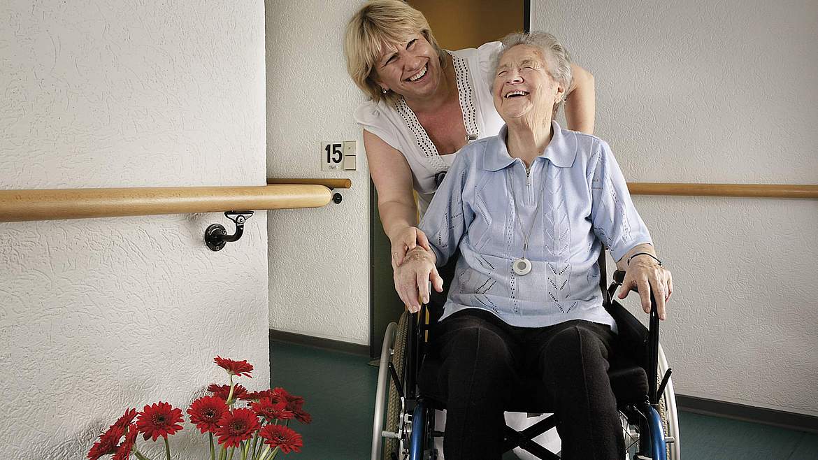 Gut betreut im Johanneswerk: Pflegekraft begleitet lachende ältere Dame im Rollstuhl