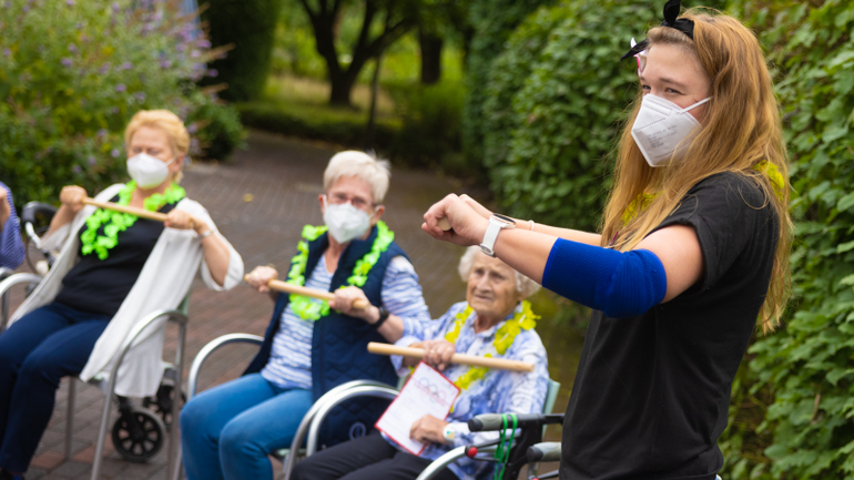 Bewegungsrunde bei der Augenblick-Olympiade Malina Sander steht bei einer Gruppe von sitzenden Menschen und macht Übungen mit einem Stab vor.