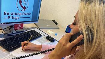 Eine blonde Frau, die vor einem PC sitzt und Telefon und einen Stift in der Hand hält