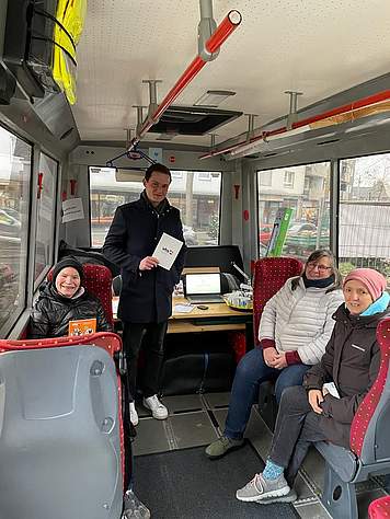 Drei Menschen sitzen in einem Bus, ein Mann steht daneben und hält eine VHS-Broschüre in der Hand