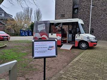 Plakat mit der Aufschrift "VHS und Jo-Mobil. Bibliothek der Digitalen Dinge" ist vor dem Jo-Mobil-Bus aufgestellt