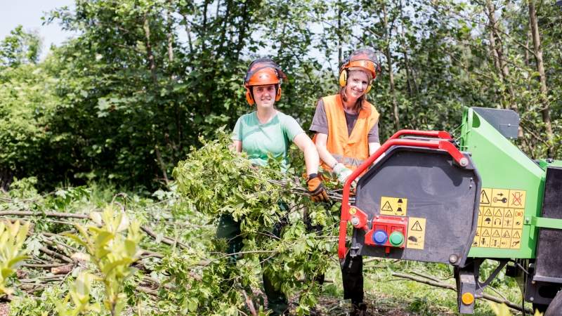 zwei Menschen mit Helmen, die an einer Maschine Holz zerkleinern