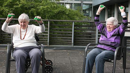 Zwei ältere Frauen sitzen auf der Terrasse und trainieren mit kleinen Sportgeräten ihre Tiefenmuskulatur.
