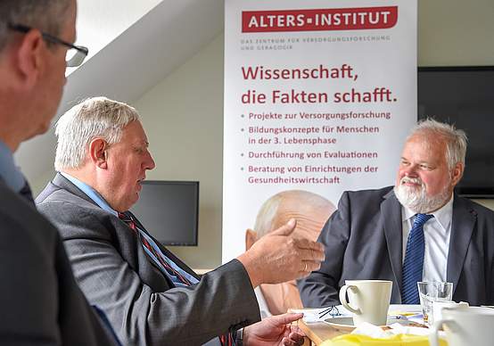 Drei Männer am Tisch unterhalten sich angeregt. Dahinter ein Schild mit der Aufschrift "Altersinstitut"