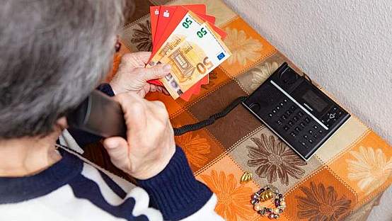 Ältere Dame mit grauem kurzen Haar und Brille von hinten, wie sie telefoniert und dabei ihr Geld und Sparbücher in der Hand hält.