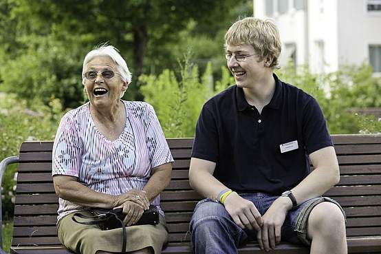 Alte Damen und junger Mann sitzen zusammen draußen im Grünen auf einer Bank und lachen gemeinsam.