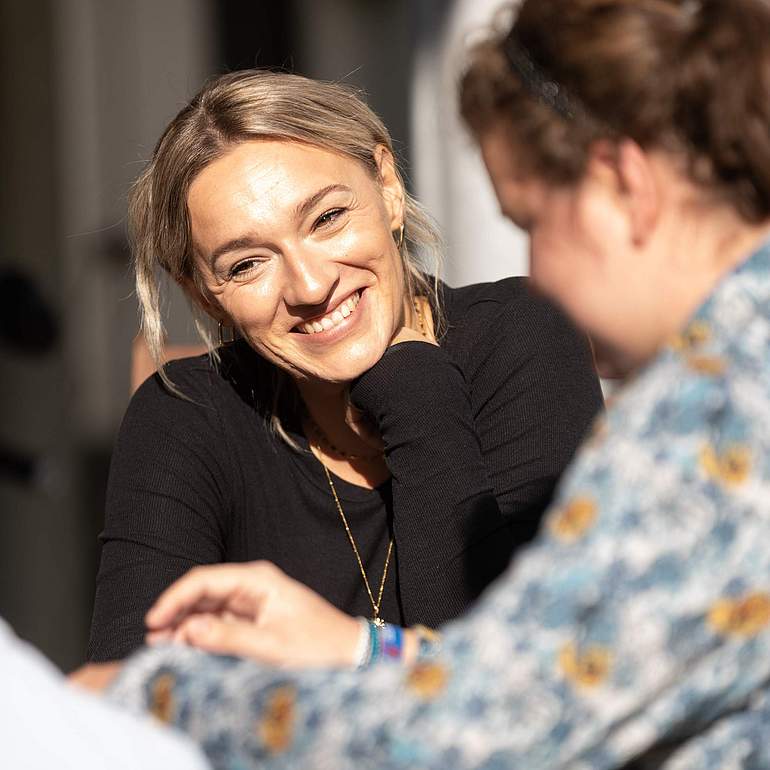 Eine blonde und eine dunkelhaarige Frau sitzen zusammen am Tisch und sprechen miteinander. 