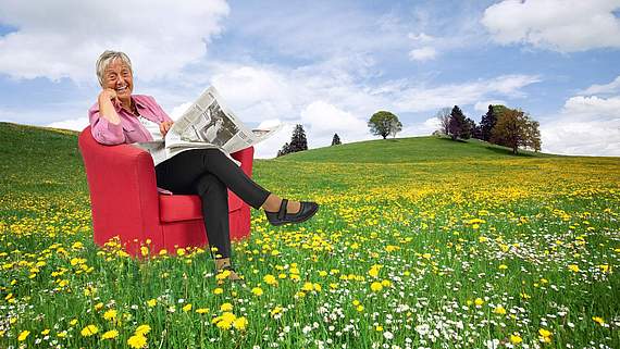 Ältere Dame mit einer Zeitung sitzt in einem roten Sessel auf einer grünen Wiese