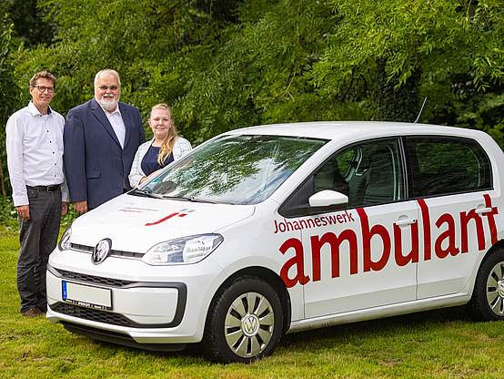 Zwei Männer und eine Frau stehen hinter einem weißen Kleinwagen und blicken in die Kamera.