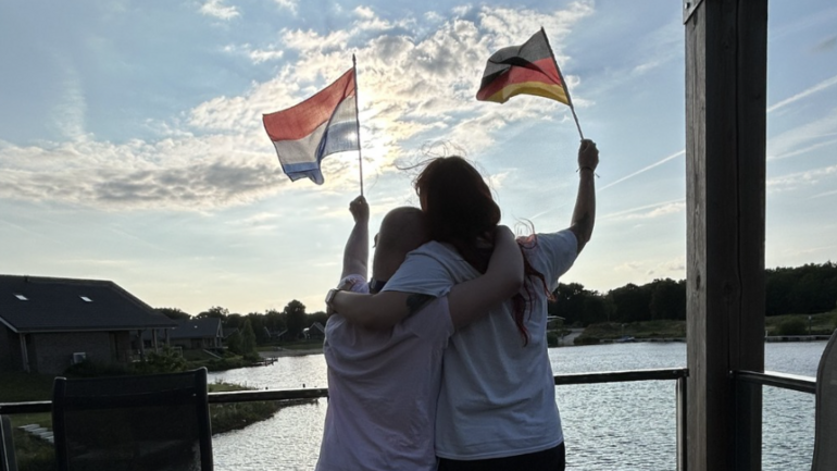 Zwei Personen stehen vor einem See und halten eine Deutschlandflagge und eine der Niederlande in den Händen
