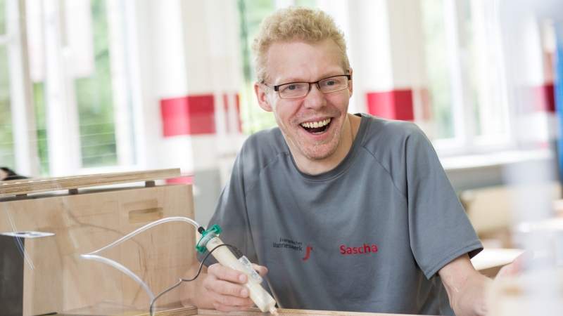 ein Mann mit grauem T-Shirt und Brille, der einen Lötkolben in der Hand hält und lächelt