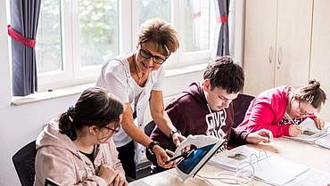 Der Berufsbildungsbereich bekommt eine neue fachliche Ausrichtung drei Personen die an einem Tisch sitzen. Eine Frau steht dahinter und erklärt ein Bügeleisen