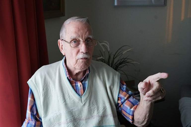 Bewohner Hans Bernoth Ein Mann sitzt in seinem Zimmer und zeigt mit seiner linken Hand zur Seite