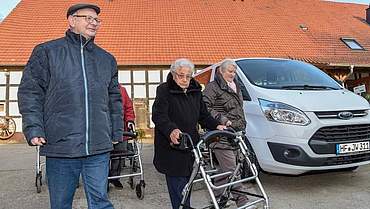 Ein Mann und zwei Frauen - davon eine mit Rollator - neben einem Bulli