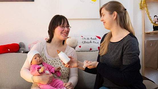 Frau hält einen Babysimulator auf dem Arm und unterhält sich mit einer anderen Frau.